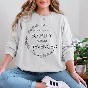 Op de afbeelding: Een grijs sweatshirt met een bloemenkransontwerp en de tekst "Wees blij dat we gelijkheid willen en geen wraak".