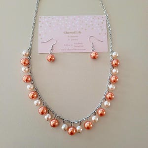 May include: A silver chain necklace with peach and white pearl beads. Matching peach pearl earrings are included. The card reads "CharmLife Accessories & Jewelry Follow us Facebook Instagram www.charmilife.etsy.com"