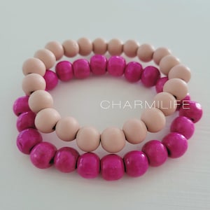 May include: Two stacked bracelets made of wooden beads. The top bracelet is made of light brown beads and the bottom bracelet is made of bright pink beads.