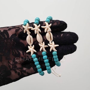 May include: Three turquoise blue beaded bracelets with white starfish and shell charms. The bracelets are stacked on top of each other and are tied with a white string.