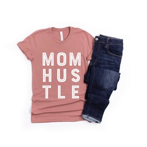 Pu&ograve; includere: Una maglietta color malva con il testo "MOM HUSTLE" in lettere bianche con contorno. La maglietta &egrave; abbinata a un paio di jeans blu scuro.
