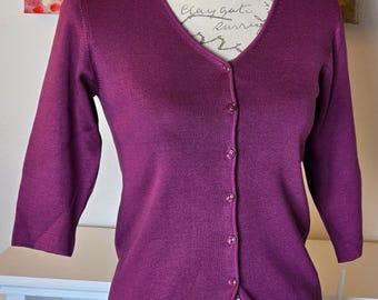 May include: A plum-colored cardigan with a V-neck and three-quarter sleeves. The button-down sweater is displayed on a mannequin. The knitwear has a ribbed texture and is a solid color.