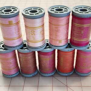 May include: A collection of pink and rose-colored thread spools, stacked on a grid surface. The spools have gray tops and bottoms, with labels and text visible. The thread colors range from light pink to deep rose, suitable for sewing and crafting projects.