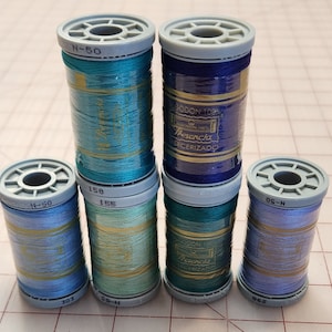May include: A collection of six spools of thread in various shades of blue, teal, and green. The spools are labeled with numbers and the brand name "Presencia". The thread is wrapped around gray plastic spools.