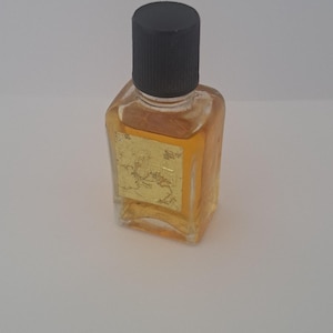 Könnte beinhalten: Eine kleine, rechteckige Glasflasche gefüllt mit einer bernsteinfarbenen Flüssigkeit. Die Flasche hat einen schwarzen Schraubverschluss und ein quadratisches, goldfarbenes Etikett auf der Vorderseite mit einer Prägung eines Schiffes.