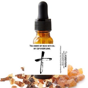May include: A small amber glass bottle with a black dropper top and a label that reads "YOU ANOINT MY HEAD WITH OIL: MY CUP OVERFLOWS." The label also features a black cross. The bottle is surrounded by small, amber-colored resin pieces.
