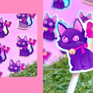 May include: Purple cat sticker with a pink bow and bell collar. The cat has large eyes and is sitting down. The sticker is on a pink background with other cat stickers. The sticker is on a white stick with green grass in the background.