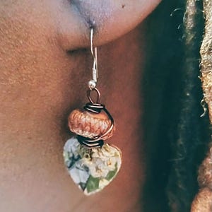 May include: Handmade earrings with a unique design. Each earring features a heart-shaped bead with a floral pattern, paired with a textured, round bead. The beads are connected with copper wire and hang from a silver hook.