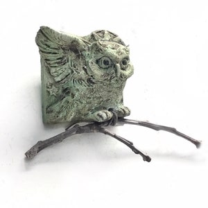 May include: A small, weathered, light green owl figurine with textured feathers and large eyes, perched on a thin, dark brown twig. The owl is attached to a square base featuring a star and circular pattern, creating a detailed, sculptural piece.