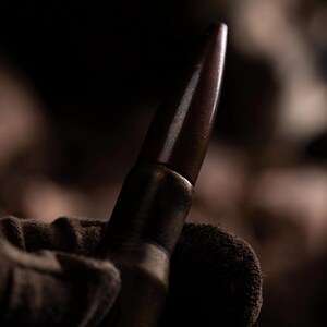 May include: A metallic cartridge featuring a pointed, dark brown projectile tip and a cylindrical brass casing. The object is held in a textured, dark-colored fabric material, with the metallic surface reflecting light along its tapered shape.
