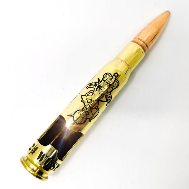 May include: A polished brass bullet casing bottle opener with a copper-colored tip. The casing features a black engraved emblem of a crown and horse with the letters R.E.M.E. and the text 24 WHISPER stamped near the base of the metal body.