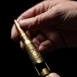 May include: A gold-colored metal bottle opener shaped like a large caliber bullet casing. The casing features an engraved emblem with a crown and the text "PER MARE PER TERRAM" visible on the side, with a cutout section for opening bottles.