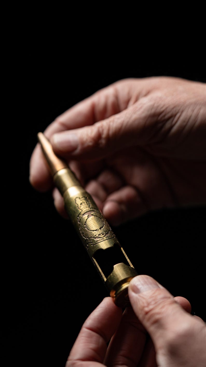 May include: A gold-colored metal bottle opener shaped like a large caliber bullet casing. The casing features an engraved emblem with a crown and the text "PER MARE PER TERRAM" visible on the side, with a cutout section for opening bottles.