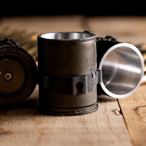 Pode incluir: Um conjunto de copos de shot de metal verde-oliva feitos a partir de carcaças de granadas reaproveitadas. Cada recipiente cilíndrico apresenta uma borda interna metálica e texto gravado na base "GRENADE CASING BY CUSTOM CALIBER".