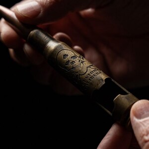 May include: A metallic, brass-colored bottle opener shaped like a large caliber bullet casing. The surface features an engraved skull and crossbones design along with the text "CUSTOM FIBERS" etched into the side of the cylindrical metal body.