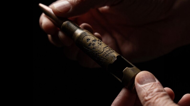 May include: A metallic, brass-colored bottle opener shaped like a large caliber bullet casing. The surface features an engraved skull and crossbones design along with the text "CUSTOM FIBERS" etched into the side of the cylindrical metal body.