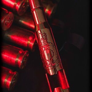 May include: A metallic bottle opener fashioned from a large caliber bullet casing, featuring an engraved emblem with the text Gibraltar and Mare Per Terram, positioned among several other brass bullet casings under red lighting.