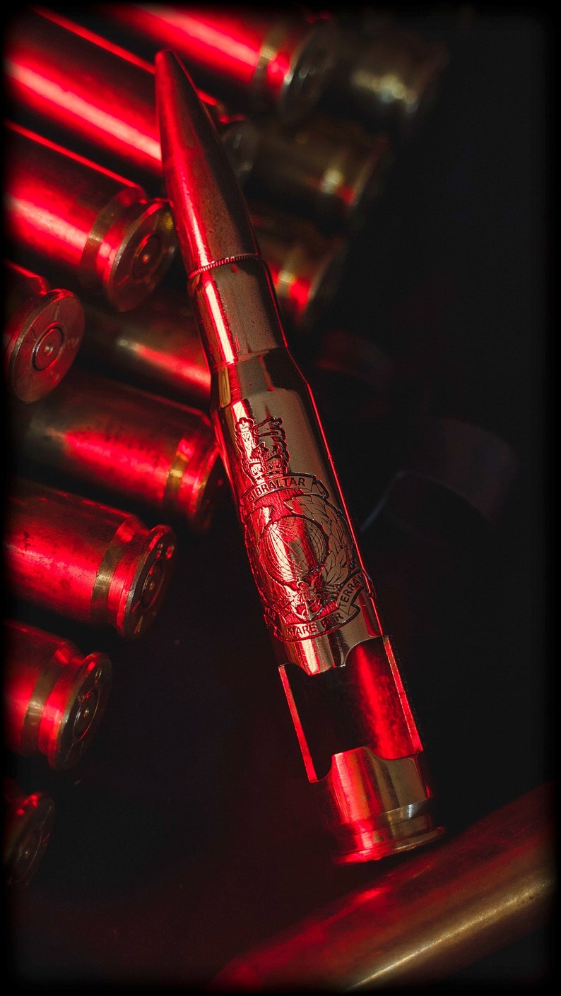 May include: A metallic bottle opener fashioned from a large caliber bullet casing, featuring an engraved emblem with the text Gibraltar and Mare Per Terram, positioned among several other brass bullet casings under red lighting.