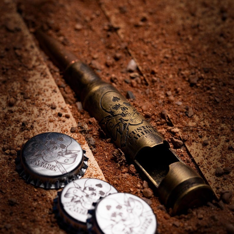 May include: A brass-colored bottle opener shaped like a large caliber bullet casing, featuring an engraved skull and crossbones design with the text CUSTOM CALIBERS. Three silver-colored metal bottle caps with matching skull graphics are included.