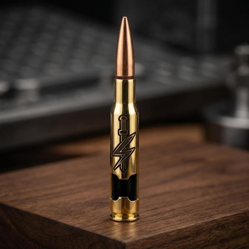 May include: A gold-colored metal bottle opener shaped like a large caliber bullet. The casing features a dark engraved design of a vertical sword overlaid with a lightning bolt. The tip of the bullet is copper-toned, and the base is brass-colored.
