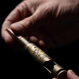 May include: A brass-colored bottle opener fashioned from a large caliber bullet casing. The metal surface features an engraved skull and crossbones graphic along with the text CUSTOM CALIBER. The object has a pointed tip and a cut-out opening.