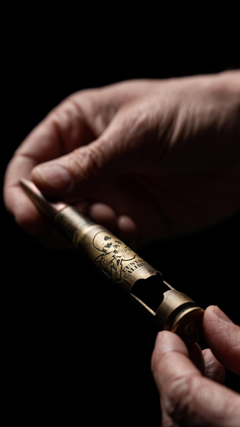 May include: A brass-colored bottle opener fashioned from a large caliber bullet casing. The metal surface features an engraved skull and crossbones graphic along with the text CUSTOM CALIBER. The object has a pointed tip and a cut-out opening.