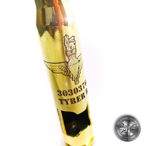 May include: A polished gold-colored metal bottle opener crafted from a large caliber shell casing. The surface features an engraved parachute emblem with a crown above it, the number 3030376, the name TYRER, and the text CUSTOM CALIBERS.