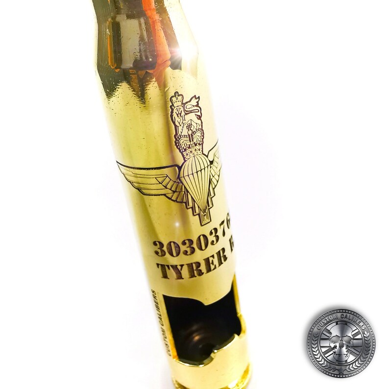 May include: A polished gold-colored metal bottle opener crafted from a large caliber shell casing. The surface features an engraved parachute emblem with a crown above it, the number 3030376, the name TYRER, and the text CUSTOM CALIBERS.