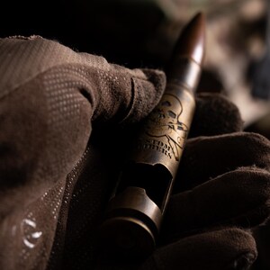May include: A brass-colored metal bottle opener shaped like a large caliber bullet casing. The casing features an engraved skull and crossbones design above the text CUSTOMERS. The opener has a rectangular cutout on the side for bottle caps.
