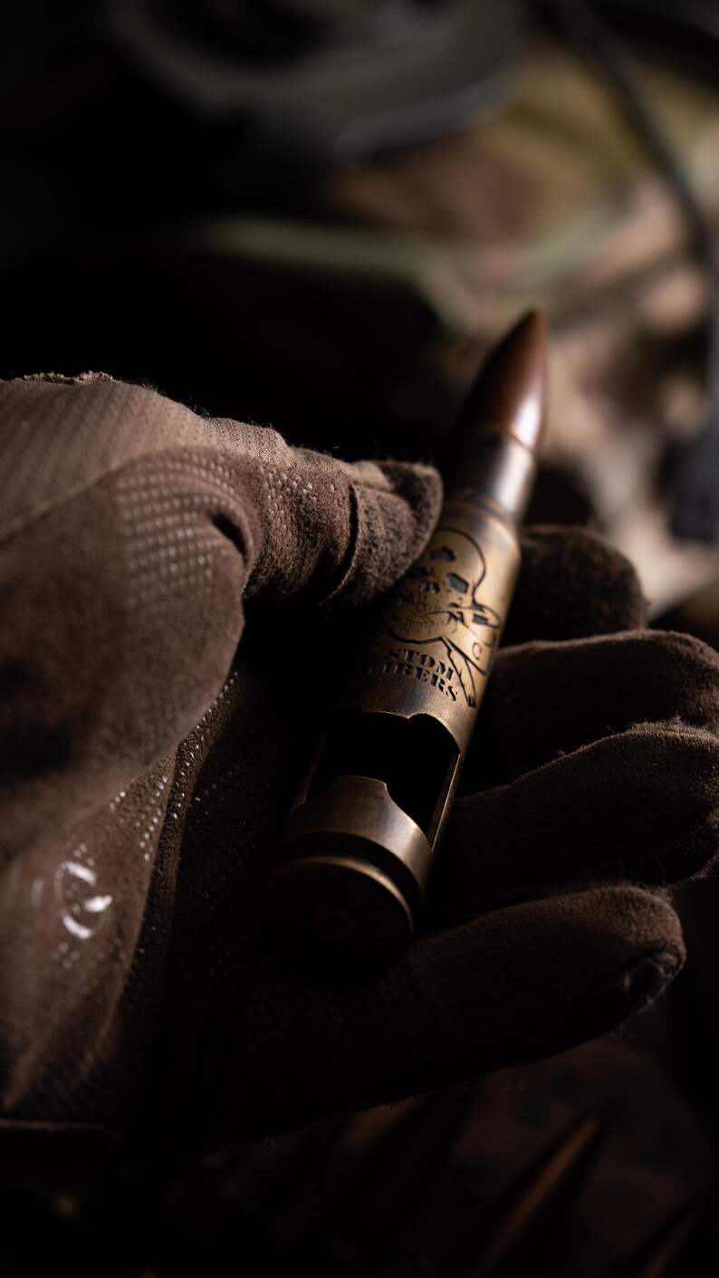 May include: A brass-colored metal bottle opener shaped like a large caliber bullet casing. The casing features an engraved skull and crossbones design above the text CUSTOMERS. The opener has a rectangular cutout on the side for bottle caps.