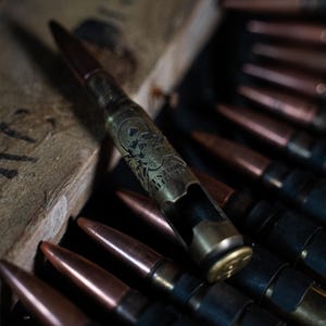 May include: A brass-colored bottle opener shaped like a large caliber bullet casing. The casing features an engraved skull and crossbones design with the text CUSTOM CALIBERS. It is positioned among several other copper-tipped bullet cartridges.