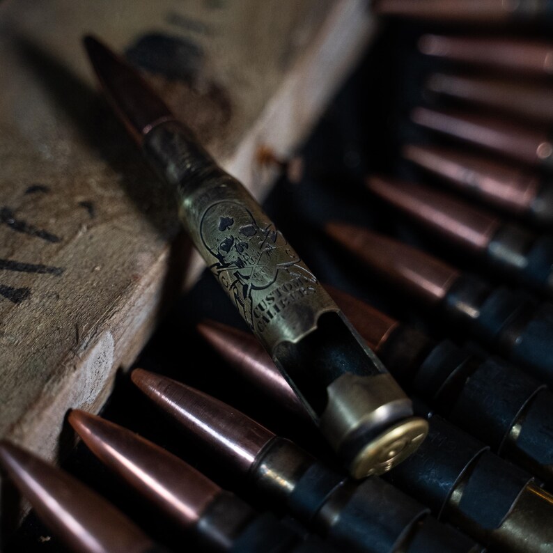 May include: A brass-colored bottle opener shaped like a large caliber bullet casing. The casing features an engraved skull and crossbones design with the text CUSTOM CALIBERS. It is positioned among several other copper-tipped bullet cartridges.