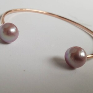 Puede incluir: Una pulsera de pu&ntilde;o de oro rosa con dos grandes perlas de color malva. La pulsera tiene un dise&ntilde;o sencillo y abierto, con las perlas situadas en cada extremo de la banda curvada.