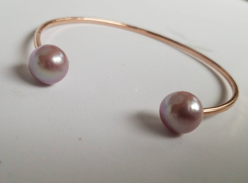 Puede incluir: Una pulsera de pu&ntilde;o de oro rosa con dos grandes perlas de color malva. La pulsera tiene un dise&ntilde;o sencillo y abierto, con las perlas situadas en cada extremo de la banda curvada.