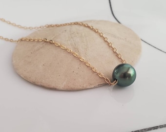 Può includere: Una collana a catena color oro con un singolo pendente di perla verde scuro. La collana è appoggiata su una superficie beige strutturata. La catena è delicata e la perla è rotonda e lucente.