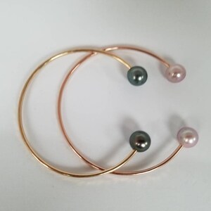 Puede incluir: Dos brazaletes abiertos de oro y oro rosa. Cada brazalete tiene una perla en cada extremo, con una perla gris oscuro y una perla rosa claro en cada brazalete. Los brazaletes est&aacute;n sobre un fondo blanco.