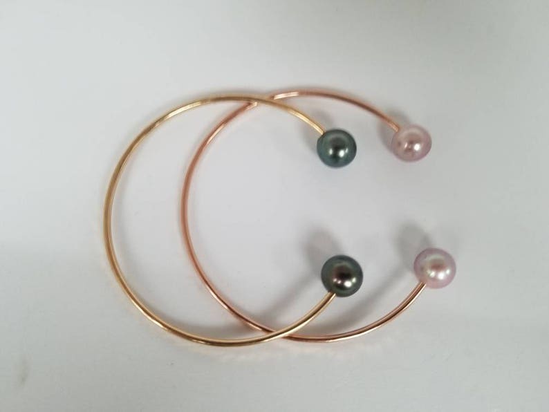 Puede incluir: Dos brazaletes abiertos de oro y oro rosa. Cada brazalete tiene una perla en cada extremo, con una perla gris oscuro y una perla rosa claro en cada brazalete. Los brazaletes est&aacute;n sobre un fondo blanco.