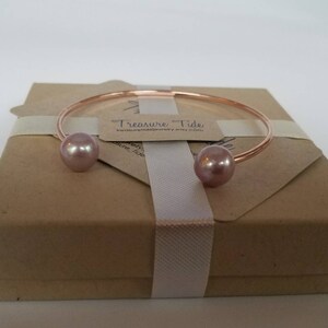 Puede incluir: Un brazalete de oro rosa con dos perlas grandes de color rosa claro. El brazalete se presenta en una caja de regalo marr&oacute;n atada con una cinta blanca. Se adjunta una peque&ntilde;a etiqueta con el texto "Treasure Tide".