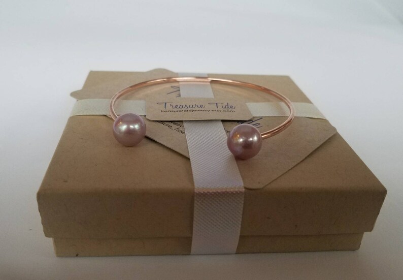 Puede incluir: Un brazalete de oro rosa con dos perlas grandes de color rosa claro. El brazalete se presenta en una caja de regalo marr&oacute;n atada con una cinta blanca. Se adjunta una peque&ntilde;a etiqueta con el texto "Treasure Tide".