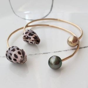Può includere: Due braccialetti dorati. Uno presenta due conchiglie con macchie nere e marroni. L'altro ha una perla dorata e una perla grigio scuro. I gioielli sono esposti su una superficie bianca.