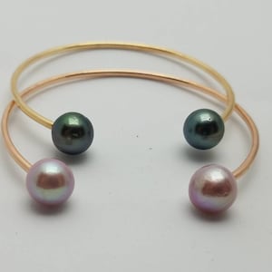 Puede incluir: Dos brazaletes abiertos, uno dorado y otro de oro rosa, cada uno con una perla brillante en cada extremo. Un brazalete tiene perlas verde oscuro, el otro perlas rosas. La joyer&iacute;a est&aacute; sobre un fondo blanco.