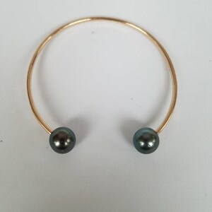 Puede incluir: Un brazalete abierto de color dorado con una perla gris oscuro en cada extremo. El brazalete tiene un dise&ntilde;o simple y elegante, perfecto para el uso diario o ocasiones especiales. Las perlas tienen un brillo sutil.