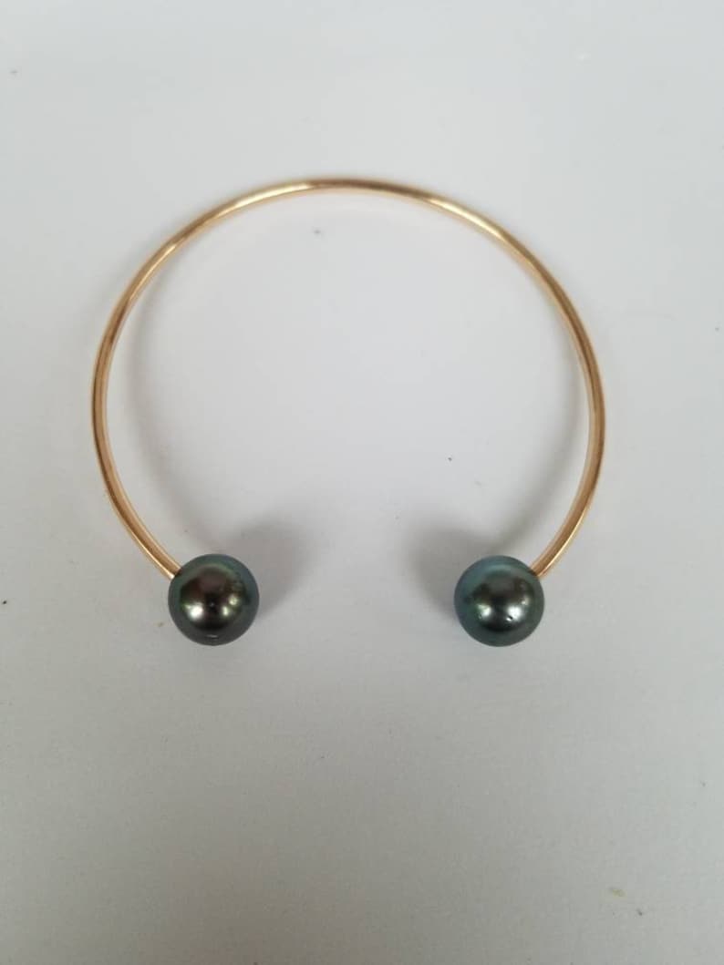 Puede incluir: Un brazalete abierto de color dorado con una perla gris oscuro en cada extremo. El brazalete tiene un dise&ntilde;o simple y elegante, perfecto para el uso diario o ocasiones especiales. Las perlas tienen un brillo sutil.