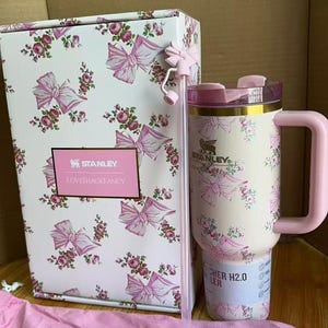 Puede incluir: Un vaso Stanley x LoveShackFancy con estampado de flores y lazos, asa rosa, tapa transparente y pajita a juego con un adorno de lazo. El vaso se muestra junto a su caja estampada a juego y una bolsa de tela rosa con texto de la marca.