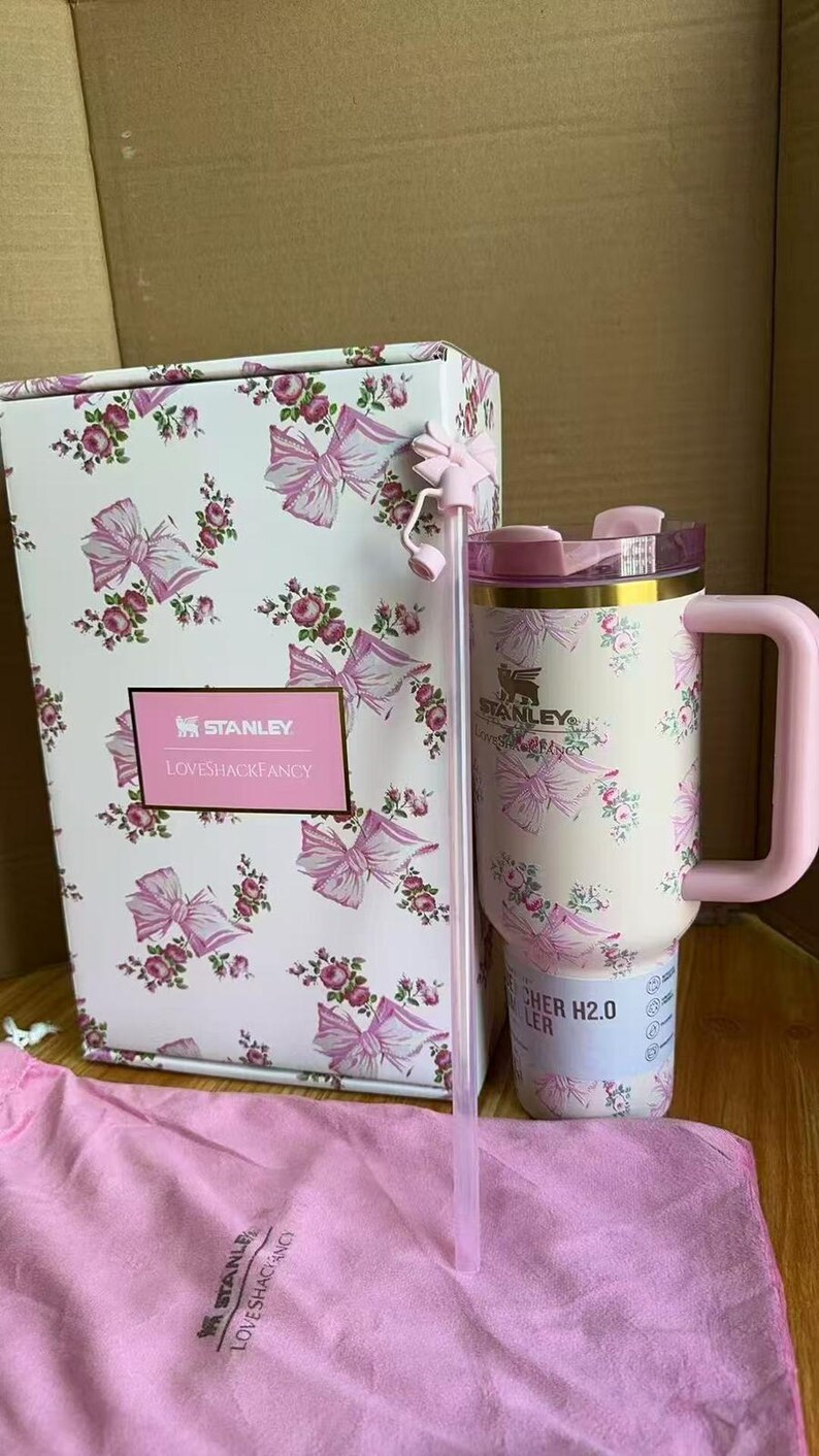 Puede incluir: Un vaso Stanley x LoveShackFancy con estampado de flores y lazos, asa rosa, tapa transparente y pajita a juego con un adorno de lazo. El vaso se muestra junto a su caja estampada a juego y una bolsa de tela rosa con texto de la marca.
