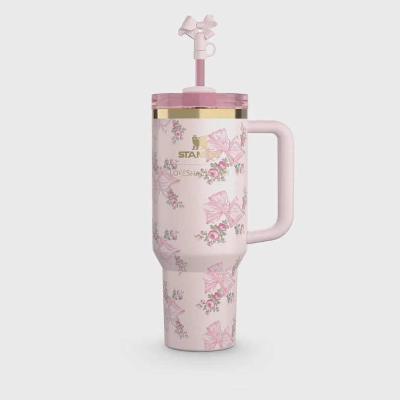 Puede incluir: Un vaso t&eacute;rmico de color rosa claro con asa, que presenta un patr&oacute;n repetitivo de lazos rosas y peque&ntilde;os grupos de flores. El logotipo de Stanley y el texto LoveShackFancy est&aacute;n impresos en dorado. Incluye una pajita rosa a juego con un adorno de lazo.