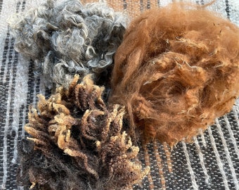 May include: Three distinct clusters of raw, unspun animal fiber. One cluster is a curly, light gray wool. Another is a dense, fluffy, reddish-brown mass of fiber. The third is a dark brown, tightly coiled, and textured clump of raw wool.