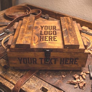 May include: A rectangular wooden crate with a dark brown stained finish, featuring two rope handles on the sides and a metal latch. The lid is engraved with the text YOUR LOGO HERE, and the front panel is engraved with the text YOUR TEXT HERE.