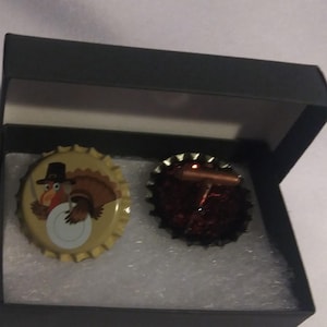 May include: A pair of cufflinks in a black box. One cufflink is a bottle cap with a cartoon turkey wearing a pilgrim hat. The other cufflink is a dark red, textured design with a copper-colored bar.