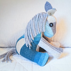 Può includere: Un giocattolo a forma di cavallo di peluche fatto a mano nei toni del blu, grigio e crema. Il cavallo ha una lunga criniera e coda grigie, con un piccolo occhio a bottone nero. Il giocattolo è appoggiato su una superficie bianca, con un cuscino a righe sullo sfondo.